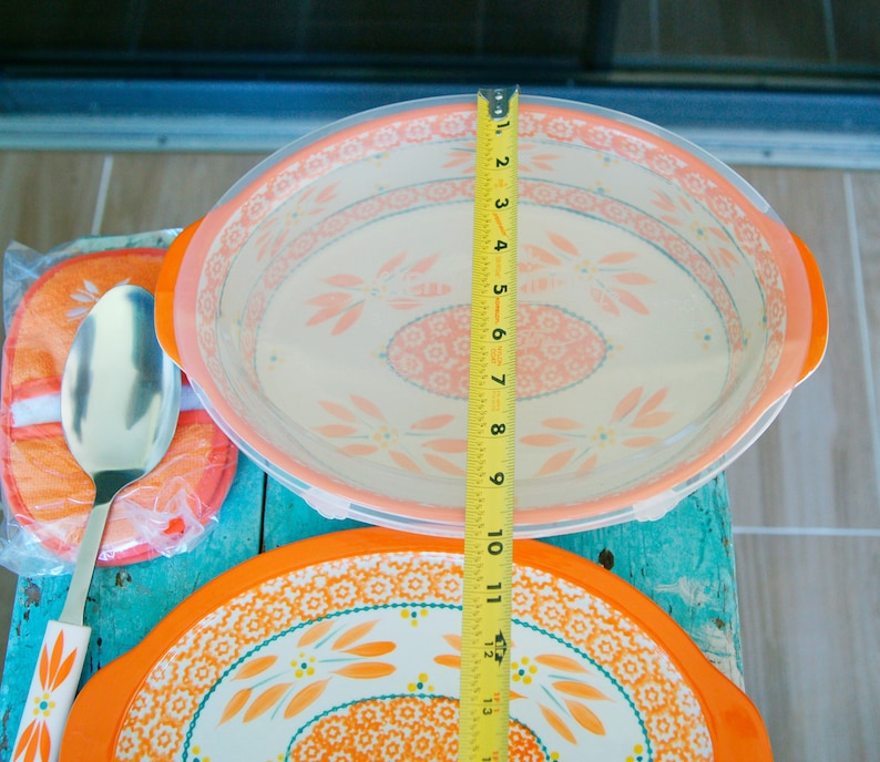 May include: An oval serving dish with a clear lid, orange handles, and a floral design. The dish measures approximately 8 inches in diameter. A matching spoon and trivet are also visible.