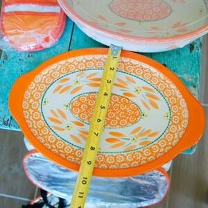 May include: An orange and white oval serving dish with a floral design, measuring approximately 10 inches in length. The dish has an orange rim and handles. A matching oven mitt and insulated bag are also visible.