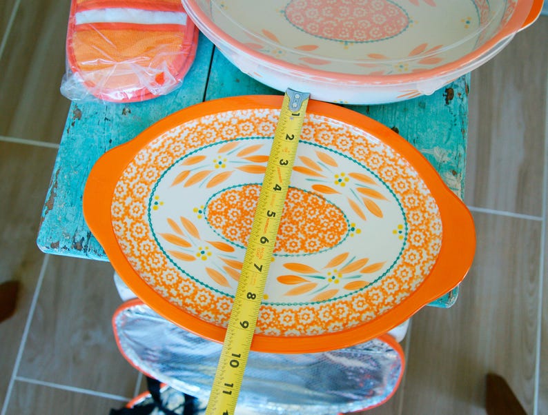 May include: An orange and white oval serving dish with a floral design, measuring approximately 10 inches in length. The dish has an orange rim and handles. A matching oven mitt and insulated bag are also visible.