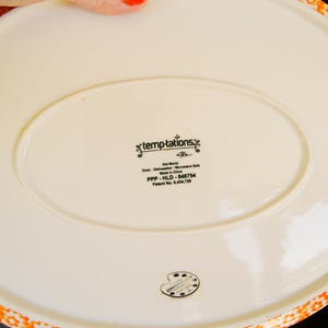 May include: Off-white oval serving platter with an orange floral patterned border. The underside features the text "temptations" and "Old World". Dishwasher and microwave safe.