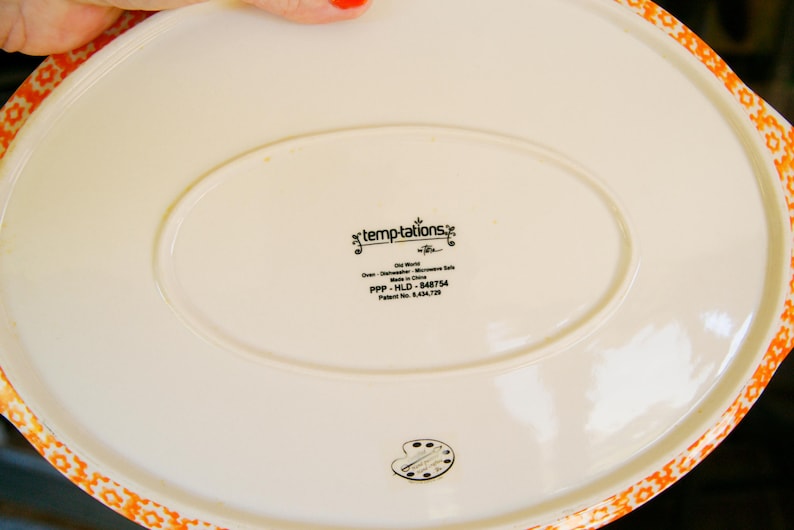 May include: Off-white oval serving platter with an orange floral patterned border. The underside features the text "temptations" and "Old World". Dishwasher and microwave safe.