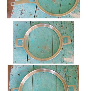 May include: A set of three stainless steel cooking pans. One pan has a long handle, while the other two have two small handles. The pans are circular and have a silver finish. The background is a distressed, turquoise-colored wooden surface.