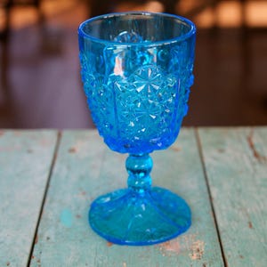 May include: A vibrant blue glass goblet with intricate embossed patterns. The stemware features a detailed design on the bowl and stem, resting on a circular base. The goblet is made of transparent glass and sits on a weathered, turquoise wooden surface.