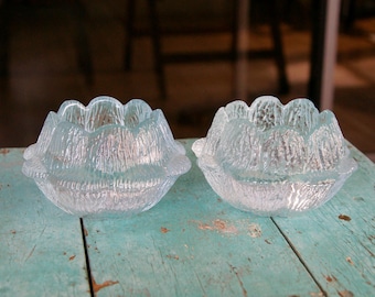 May include: Two clear glass candle holders with a textured, floral design. The candle holders are sitting on a weathered, turquoise-colored wooden surface. The candle holders have a scalloped rim.