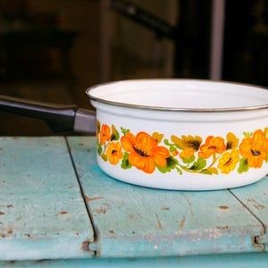May include: White enamel saucepan with a black handle and a floral design featuring orange and yellow flowers on a white background.