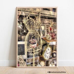 May include: A vintage-style white metal dress form with a clock and other antique items in the background. The dress form is in a shop window with a wooden frame.