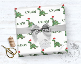 May include: White wrapping paper featuring a repeating pattern of green dinosaurs wearing Santa hats and Christmas lights, with the name "COLEMAN" in green. A silver ribbon and tag with the text "Mattie V Designs" are on top.