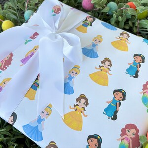 Pu&ograve; includere: Carta da regalo bianca con un fiocco di raso bianco. La carta presenta un motivo ripetuto di principesse Disney nei loro abiti caratteristici. Le principesse includono Cenerentola, Belle, Ariel, Jasmine e Rapunzel.