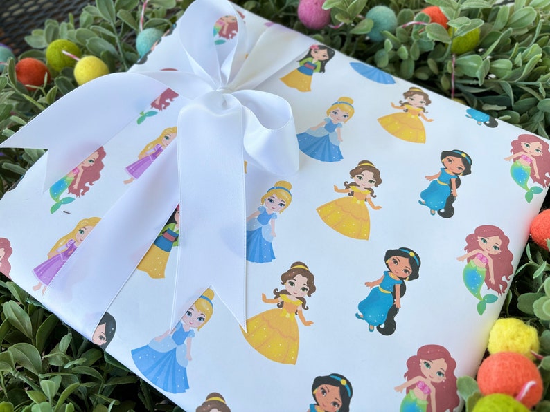 Pu&ograve; includere: Carta da regalo bianca con un fiocco di raso bianco. La carta presenta un motivo ripetuto di principesse Disney nei loro abiti caratteristici. Le principesse includono Cenerentola, Belle, Ariel, Jasmine e Rapunzel.