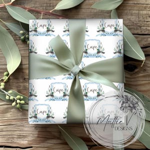 May include: White gift wrap with a repeating pattern of ducks and reeds in a blue and green watercolor design. The text "Cape" is repeated throughout the pattern. The gift wrap is tied with a sage green ribbon.