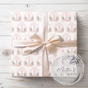 May include: Pink and white wrapping paper with a repeating pattern of a nutcracker, a Christmas tree, and a pink letter S. The paper is wrapped around a gift box with a beige ribbon.