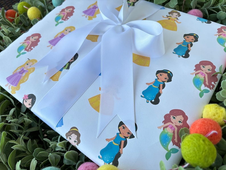 Pu&ograve; includere: Carta da regalo bianca con un fiocco di raso bianco. La carta presenta un motivo ripetuto di principesse Disney, tra cui Ariel, Belle, Jasmine e Rapunzel.
