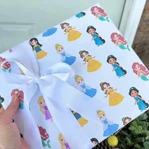 Pu&ograve; includere: Carta da regalo bianca con un motivo ripetitivo di principesse e sirene Disney. La carta &egrave; legata con un nastro di raso bianco.