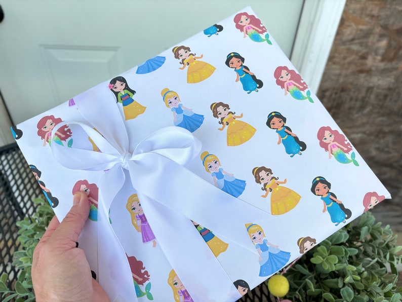 Pu&ograve; includere: Carta da regalo bianca con un motivo ripetitivo di principesse e sirene Disney. La carta &egrave; legata con un nastro di raso bianco.