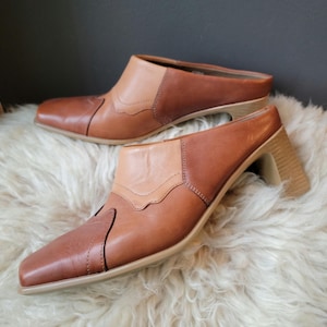 Op de afbeelding: Tweekleurige leren muiltjes met een vierkante neus en een hoge blokhak. De schoenen hebben licht- en donkerbruine leren bovenstukken met decoratieve stiksels. De hakken zijn ongeveer 7,6 cm hoog.