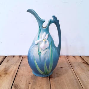 May include: A ceramic pitcher featuring a matte blue finish with a raised white iris flower and green leaves. The vessel has a curved handle and a pointed spout, showcasing a smooth, rounded body that tapers toward the top opening.