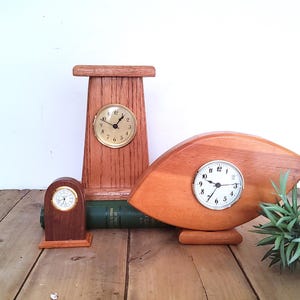 May include: Three wooden clocks of varying sizes and shapes are displayed on a rustic wooden surface. The clocks feature round faces with Roman numerals and hands. One clock is a small, arched design, another is a tall, rectangular design, and the third is a unique, curved shape.