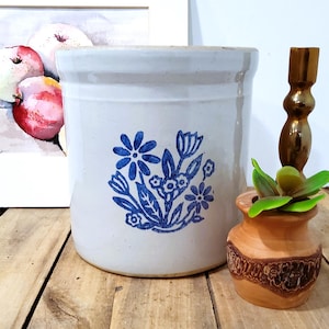 May include: A white ceramic crock with a blue floral design, a small wooden vase with a plant, a gold candlestick, and a framed watercolor painting of apples on a wooden surface.