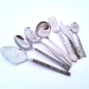 May include: A collection of seven silver-toned metal utensils including a cake server, various spoons, a fork, and knives. Each piece features a distinct handle design, ranging from smooth metallic surfaces to intricate floral and geometric patterns.