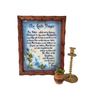 May include: A framed print of The Lord's Prayer with a floral and candle design. A brass candlestick and a small potted plant are next to the framed print. The frame is made of dark wood.
