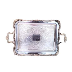 May include: A rectangular silver serving tray with ornate detailing. The tray features a floral pattern in the center and decorative handles on each side. The edges are scalloped, adding to its elegant design.