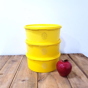 May include: Three bright yellow, round food storage containers stacked on top of each other, with a decorative floral design. A shiny red apple sits next to the containers on a wooden surface.