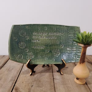 May include: A green ceramic serving platter with embossed designs and the quote "SPARROW SINGING - ITS TINY MOUTH OPEN - BUSON." The platter is displayed on a black stand. A decorative vase with a faux succulent is next to the platter.
