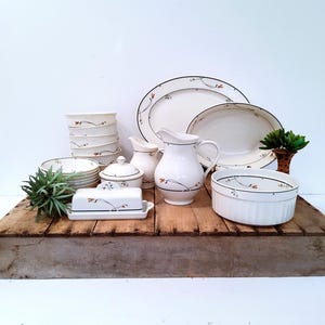 May include: A collection of white ceramic tableware with a black and red floral design. Includes plates, bowls, a pitcher, a butter dish, and a covered dish. The items are arranged on a rustic wooden surface.