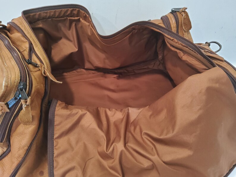 May include: Open view of a brown leather duffel bag. The bag has multiple zippered compartments and a spacious interior. The interior lining is a lighter shade of brown. The bag appears to be well-worn, with visible signs of use.