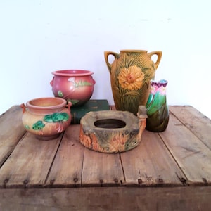 May include: A collection of five ceramic vessels including two small round pots with handles, a tall yellow vase with floral relief, a low tree-stump shaped bowl, and a small green and pink vase, all featuring textured surfaces and earthy tones.