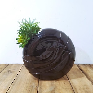 May include: A dark brown, spherical ceramic vase with a glossy finish and a ribbed design. The vase contains a small arrangement of green and yellow artificial succulents. The vase is displayed on a wooden surface.