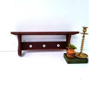 May include: A dark red wooden shelf with three white knobs below. A small terracotta pot with a plant and a brass candlestick are on the right. The shelf is designed for wall mounting.
