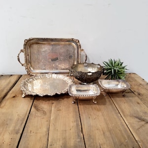 May include: A collection of silver-toned metal serving pieces including a rectangular tray with handles, a round scalloped tray, a footed bowl with ornate detailing, and two small rectangular footed dishes arranged together on a surface.