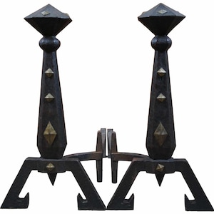 May include: Two dark metal and brass andirons with geometric shapes. Each andiron has a tall, tapered body with diamond and pyramid-shaped accents. The base has angled legs with a hook at the end.