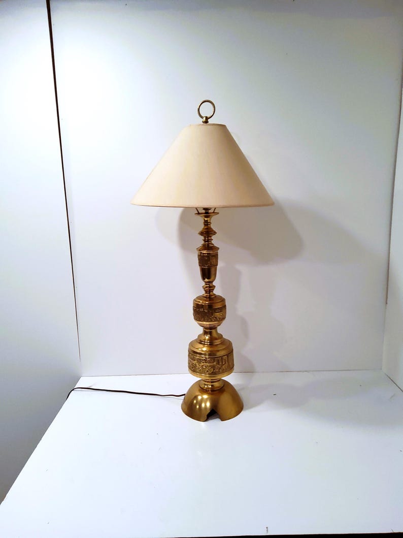 May include: A vintage brass table lamp with a cream-colored lampshade. The lamp features an ornate, detailed base and stem. The shade is conical, and the lamp has a ring pull. The lamp is approximately 28 inches tall.