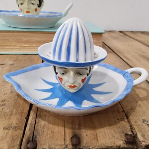May include: A white ceramic citrus juicer featuring a clown face design with red heart-shaped cheek accents and a blue star-shaped collar pattern. The juicer has a blue and white striped reamer top and a handle on the side of the base dish.