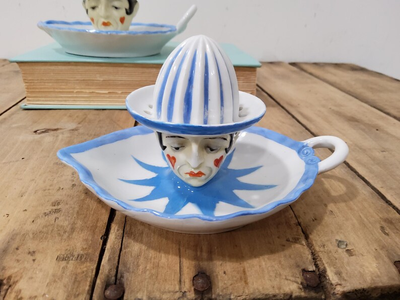 May include: A white ceramic citrus juicer featuring a clown face design with red heart-shaped cheek accents and a blue star-shaped collar pattern. The juicer has a blue and white striped reamer top and a handle on the side of the base dish.