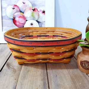 May include: Oval-shaped woven basket with a red, black, and yellow design. The basket is on a wooden surface, with a painting of apples in the background. A small decorative vase and candle holder are also present.
