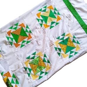 May include: White quilt with a repeating pattern of green, yellow, and orange geometric squares. The squares are set against a white background, with a green border along one edge. A handmade textile.