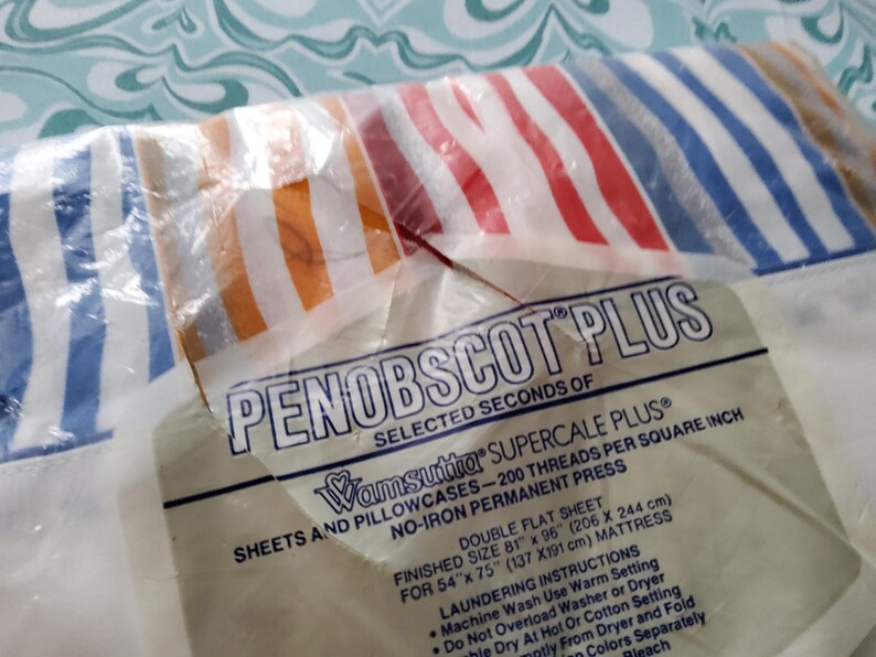 K&ouml;nnte beinhalten: Eine Packung Penobscot Plus Doppelbettlaken. Die Bettlaken haben ein gestreiftes Muster in Rot, Wei&szlig;, Blau und Orange. Die Verpackung enth&auml;lt Text &uuml;ber die Fadenzahl und die Abmessungen der Laken (206 x 244 cm).