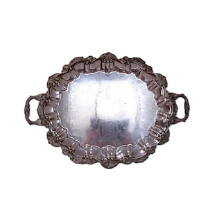May include: An oval, silver-colored serving tray with ornate detailing around the edges and two handles. The tray has a reflective surface and is set against a white background. The tray is likely made of metal.