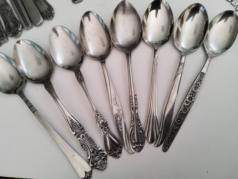 May include: A set of eight silver spoons with ornate handles. The spoons have a polished finish and are arranged in a fan shape.
