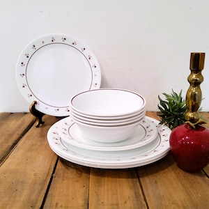 May include: A white dinnerware set with a floral pattern in burgundy. The set includes plates and bowls. A brass candlestick and a red apple-shaped ornament are also on display, creating a vintage aesthetic.