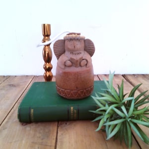 May include: A brown ceramic angel figurine with a textured surface, featuring a halo, wings, and hands holding an open book. The base of the figurine is decorated with a repeating geometric zigzag pattern carved into the clay material.