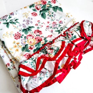 May include: White floral bedding set with a folded sheet and a ruffled edge. The floral pattern includes red, pink, and yellow roses with green leaves. The ruffled edge is trimmed with red ribbon.