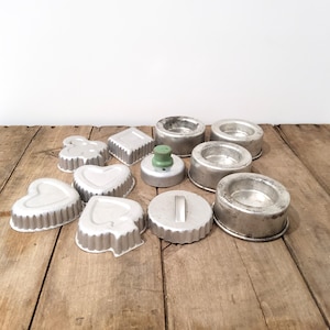 May include: A selection of vintage metal cookie cutters in diverse shapes, such as hearts, diamonds, and circles. Some cutters have fluted edges. A metal object with a green knob is also present. The items are arranged on a rustic wooden surface.