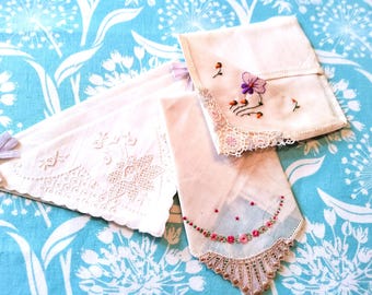 May include: Several white handkerchiefs with delicate embroidery and lace details are arranged on a blue floral patterned surface. One handkerchief features a purple flower and orange accents. Another has a floral border.