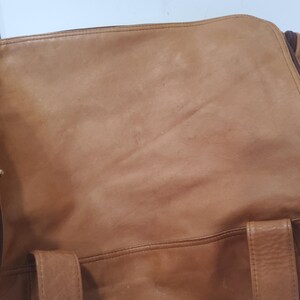May include: A tan leather duffel bag with a zipper closure and handles. The bag has a worn appearance, with visible creases and variations in color. The zipper is dark brown, and the bag appears to be well-used.