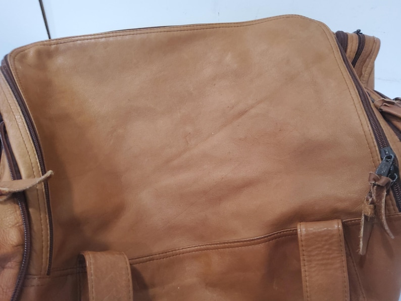 May include: A tan leather duffel bag with a zipper closure and handles. The bag has a worn appearance, with visible creases and variations in color. The zipper is dark brown, and the bag appears to be well-used.