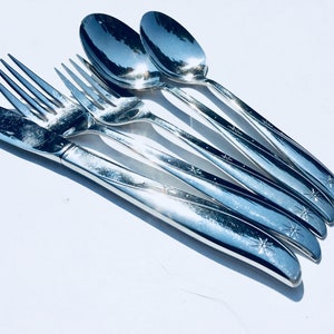 May include: A set of polished silver flatware, including a knife, forks, and spoons, arranged on a white surface. The silverware has a starburst design on the handles. The spoons reflect the surrounding environment.
